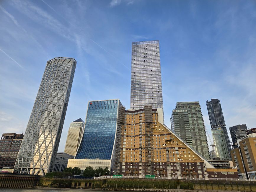 A skyline view of Canary Wharf featuring modern skyscrapers, including a tall reflective glass tower in the center, surrounded by other high-rise office buildings with varying architectural styles suc