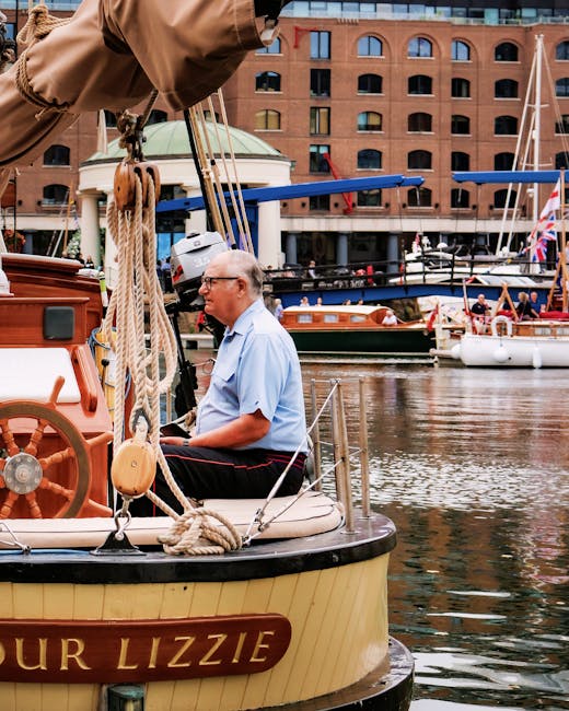 A row of moored leisure boats and yachts with white hulls and wooden decks along a calm waterway, decorated with Union Jack flags and nautical banners. The boats are situated near a walkway shaded by 